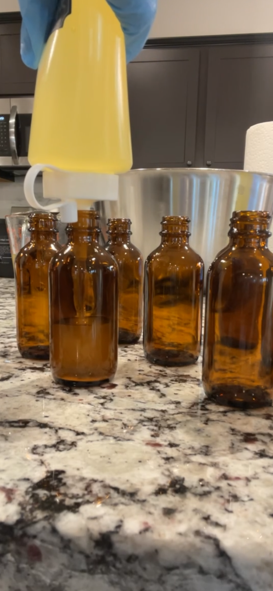 A person filling amber glass bottles with a Her Heavenly Strands Miracle Mix Hair Growth Oil using a dispenser in a kitchen.